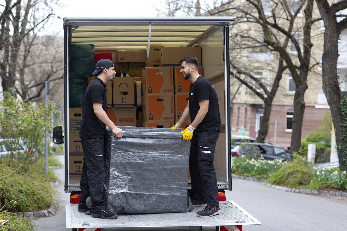 Zwei Mitarbeiter verladen einen eingepackten Sessel über die Ladebordwand in den Umzugswagen