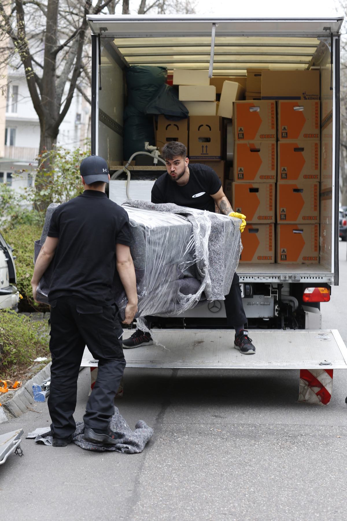 Zwei Mitarbeiter heben ein verpacktes Sofa auf die Ladebordwand des Umzugswagens